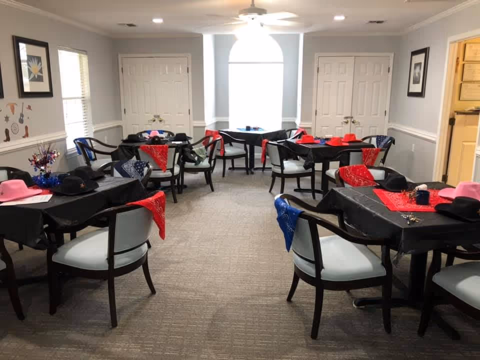 A dining room with several tables covered in black tablecloths and decorated with red, blue, and pink cowboy hats and bandanas. The room has light gray walls, white trim, and a large window with an arched top letting in natural light. Chairs with light-colored cushions surround the tables, and there are framed pictures on the walls.