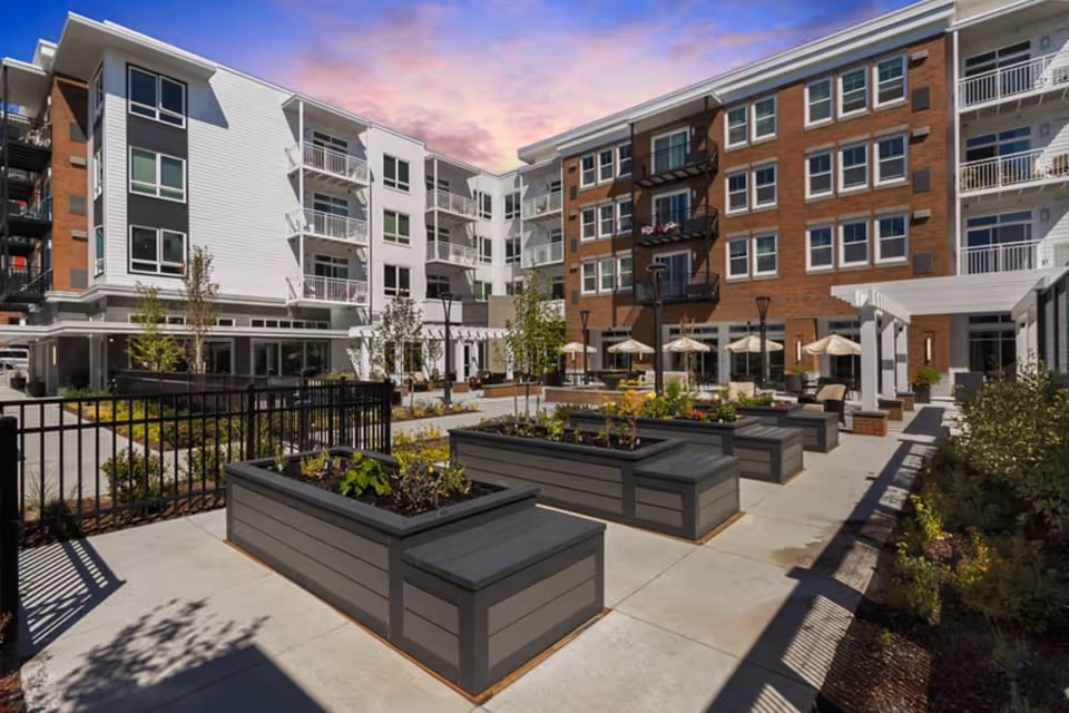 Outdoor courtyard area of a senior living facility with raised garden beds, patio seating with umbrellas, and multi-story residential buildings surrounding the space under a colorful sky at sunset.
