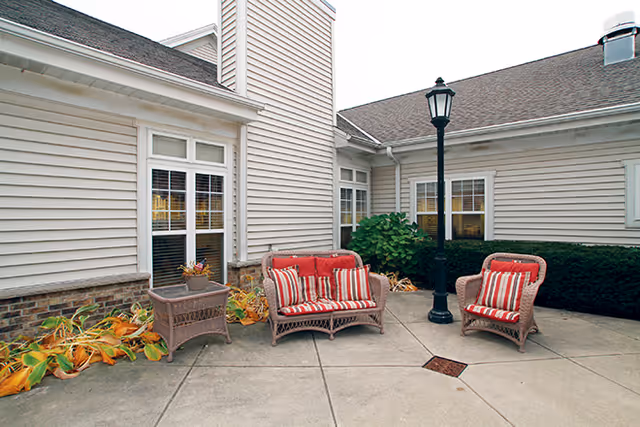 Outdoor patio area at a senior living facility with wicker furniture including a loveseat and two chairs with red and white striped cushions. The patio is surrounded by beige siding walls with windows, a black lamp post, and some plants along the edges.