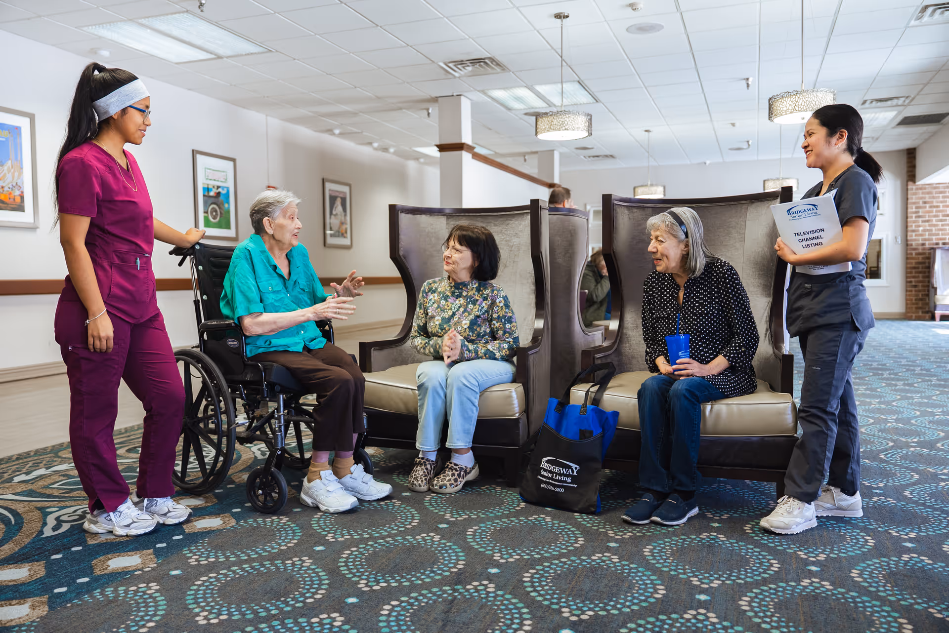 Two elderly women sitting and talking in a senior living facility common area, one in a wheelchair and the other on a chair, accompanied by two female caregivers standing nearby, one holding a folder labeled 'Bridgeway Senior Living Television Channel Listing'.