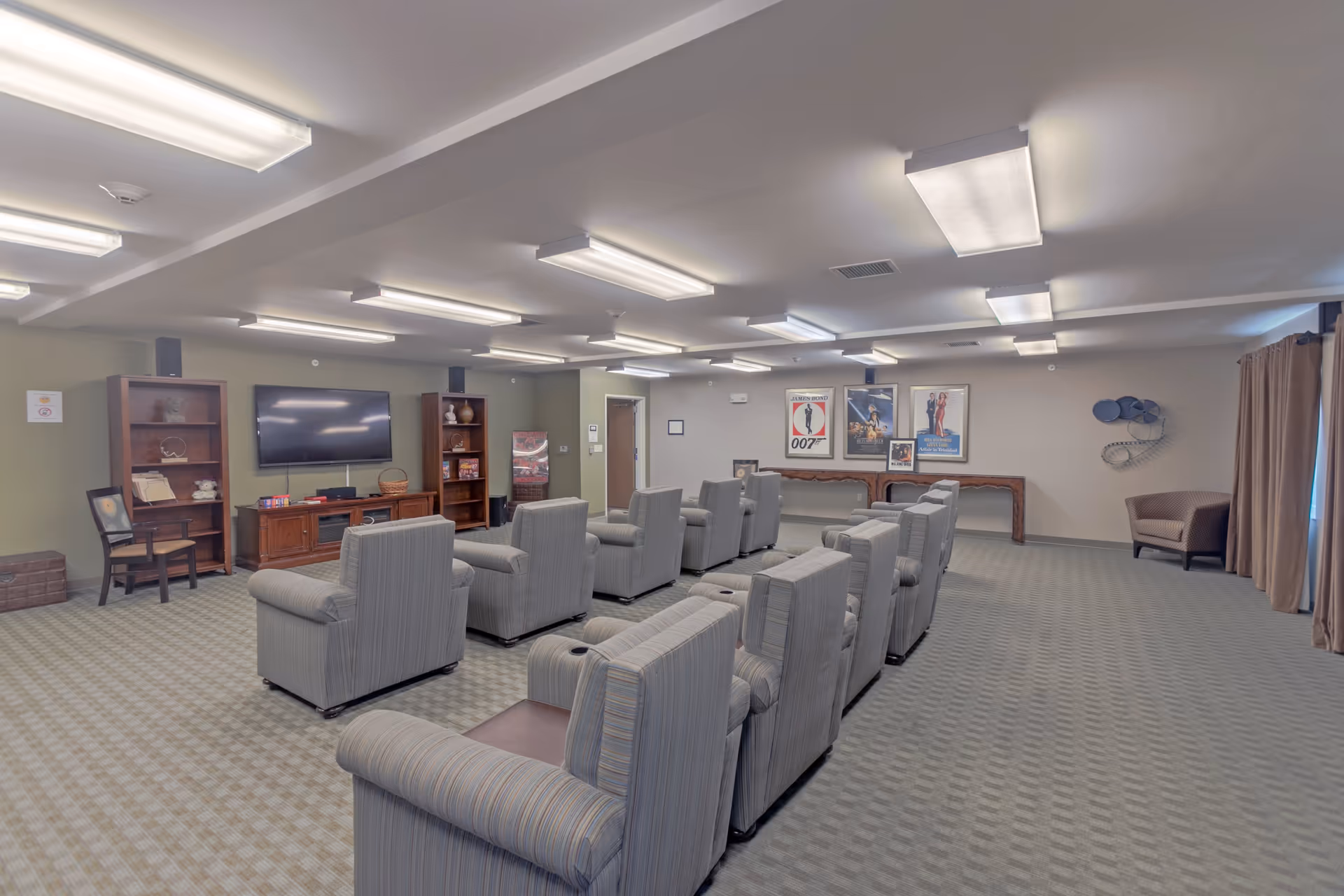A large room with multiple rows of gray upholstered armchairs facing a wall-mounted flat-screen TV. The room has carpeted floors, fluorescent ceiling lights, and bookshelves with decorative items. Movie posters are displayed on the far wall, and there is a single armchair near a window with curtains on the right side.