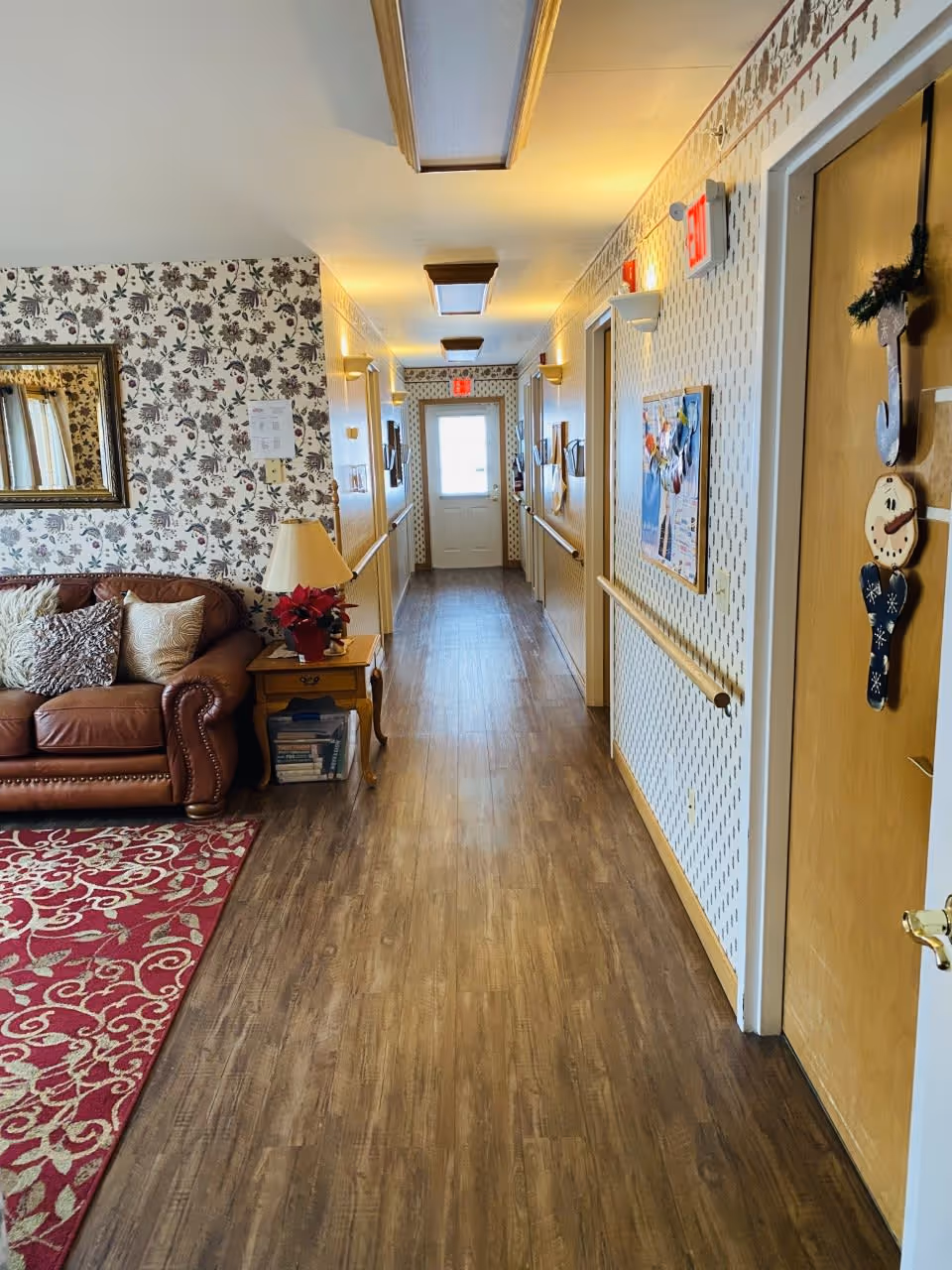 A hallway in a senior care facility with wood flooring and patterned wallpaper. On the left side, there is a brown leather couch with decorative pillows and a wooden side table with a lamp and a poinsettia plant. The hallway has handrails on both sides and several doors, one decorated with holiday ornaments. Ceiling lights illuminate the corridor, and an exit door is visible at the far end.