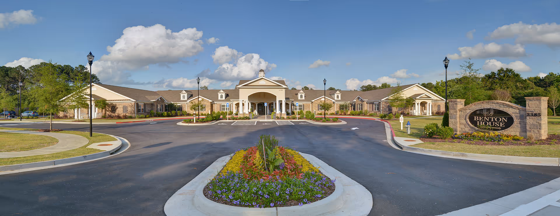Front exterior view of Benton House, a single-story building with a large covered entrance, surrounded by a circular driveway with landscaped flower beds and lamp posts under a partly cloudy blue sky.