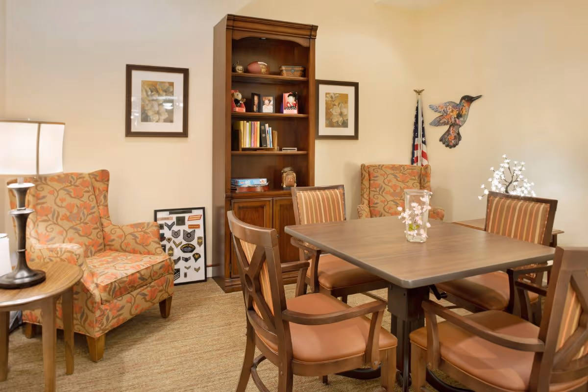 A cozy common room with a square wooden table surrounded by chairs, upholstered armchairs, and a bookcase against the wall.