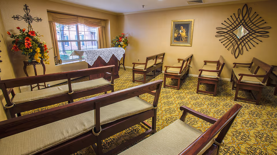 A small chapel room with wooden pews arranged in rows facing a lectern covered with a white lace cloth. The room has floral arrangements on stands near the window, a decorative cross on the wall, a framed religious painting, and a decorative mirror. The carpet has a yellow and brown intricate pattern.