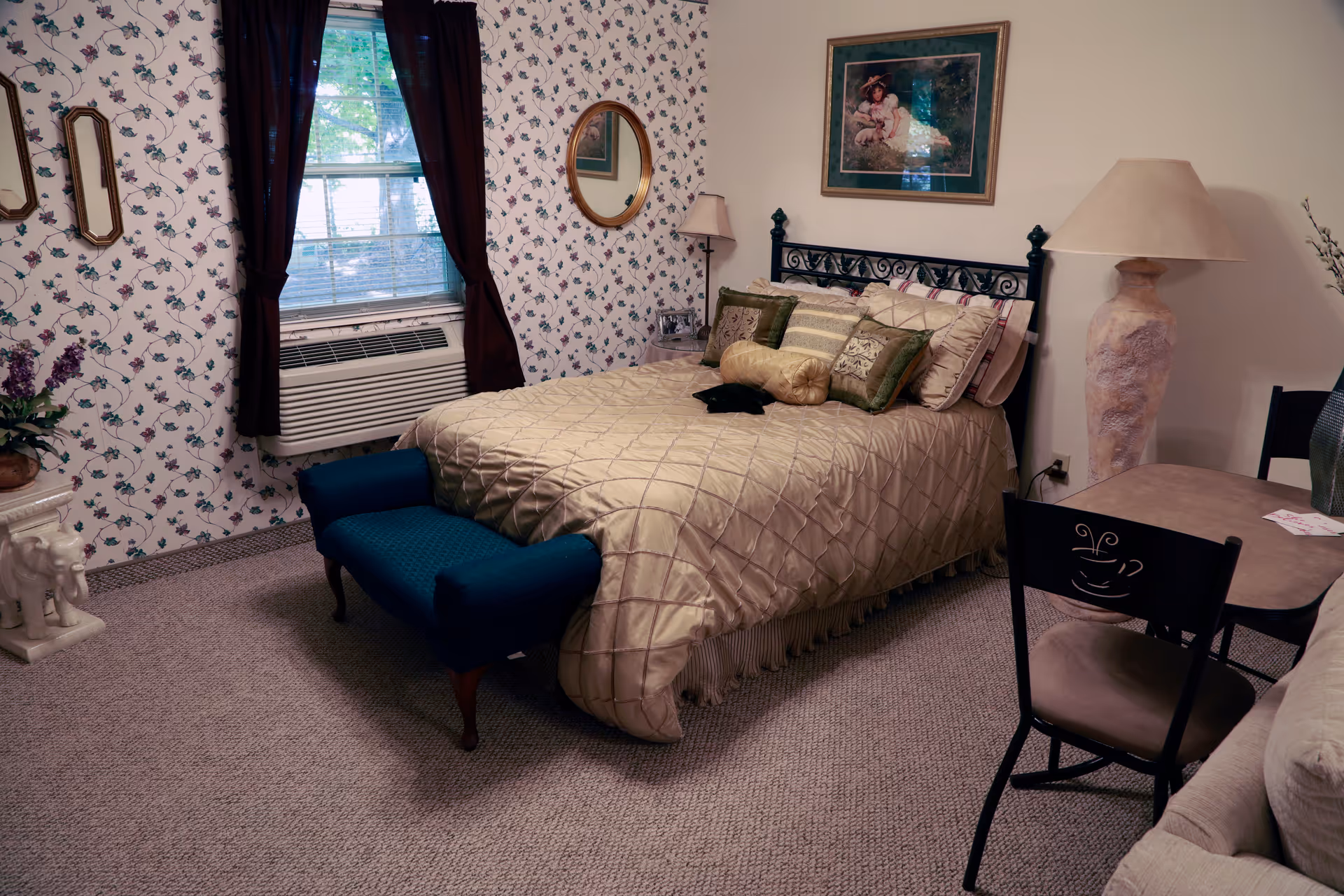 A cozy bedroom with a bed covered in a beige quilt and multiple pillows. The room has floral wallpaper on one wall, a window with dark curtains, an air conditioning unit below the window, a blue upholstered bench at the foot of the bed, a large lamp on a side table, a small round table with two chairs, and framed artwork on the wall above the bed.