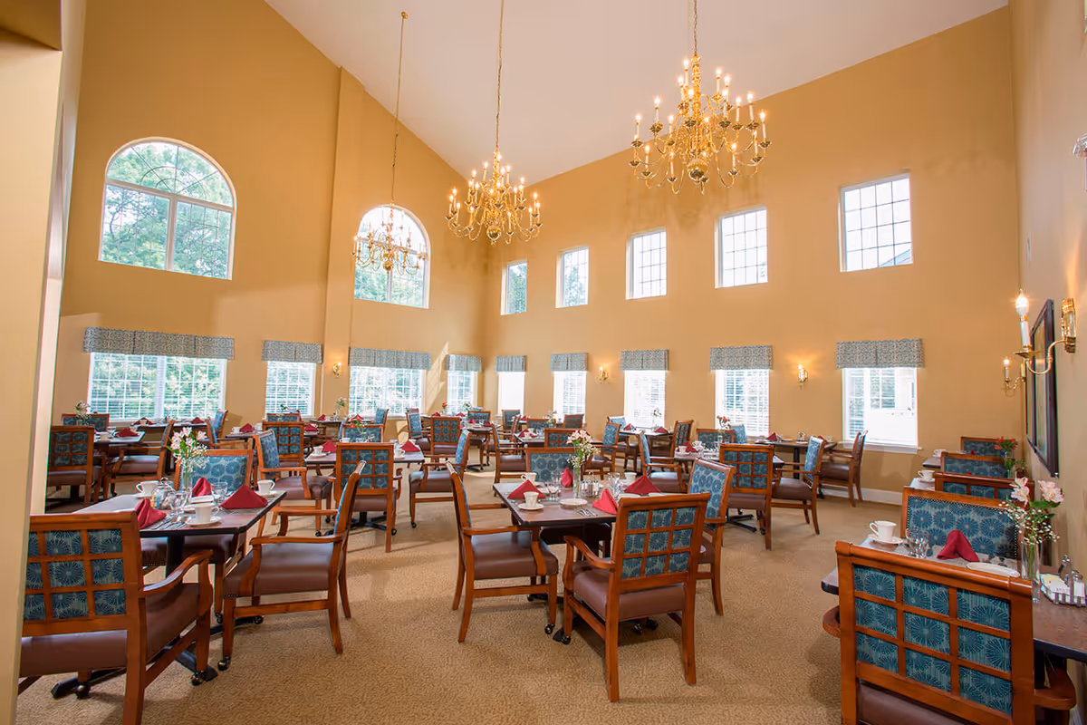 A spacious dining room with high ceilings and large windows allowing natural light to fill the space. The room features multiple wooden tables set with white cups, plates, silverware, and red folded napkins. Blue cushioned chairs with wooden frames surround each table. Elegant chandeliers hang from the ceiling, and small flower arrangements decorate the tables.