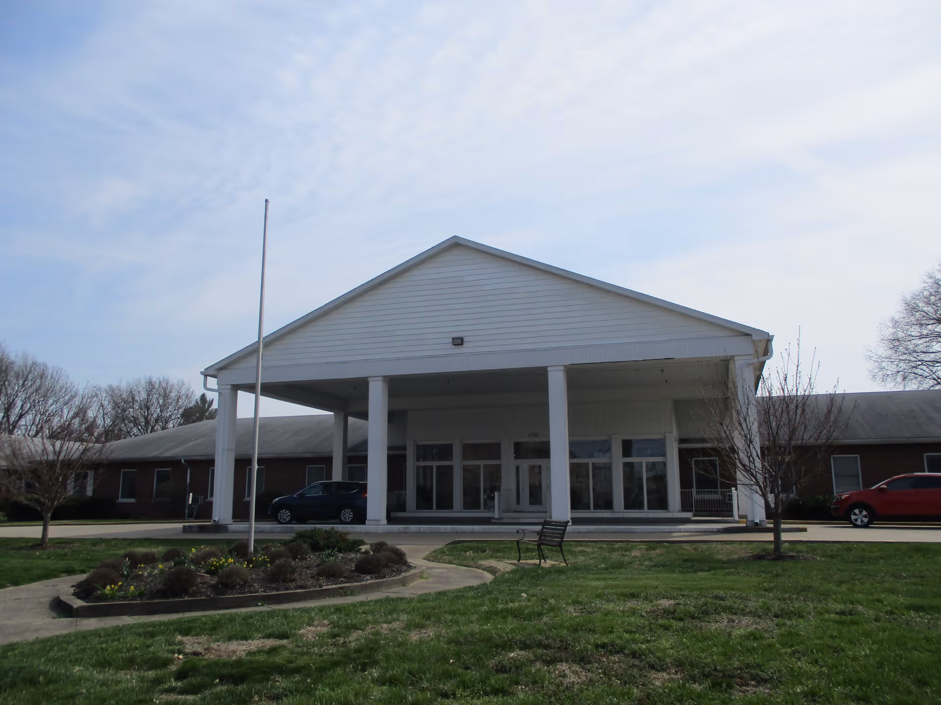 Front exterior view of a single-story building with a large covered entrance supported by white columns. There is a flagpole without a flag in front, a small landscaped garden, a bench, and two parked cars on either side of the entrance. The sky is partly cloudy.