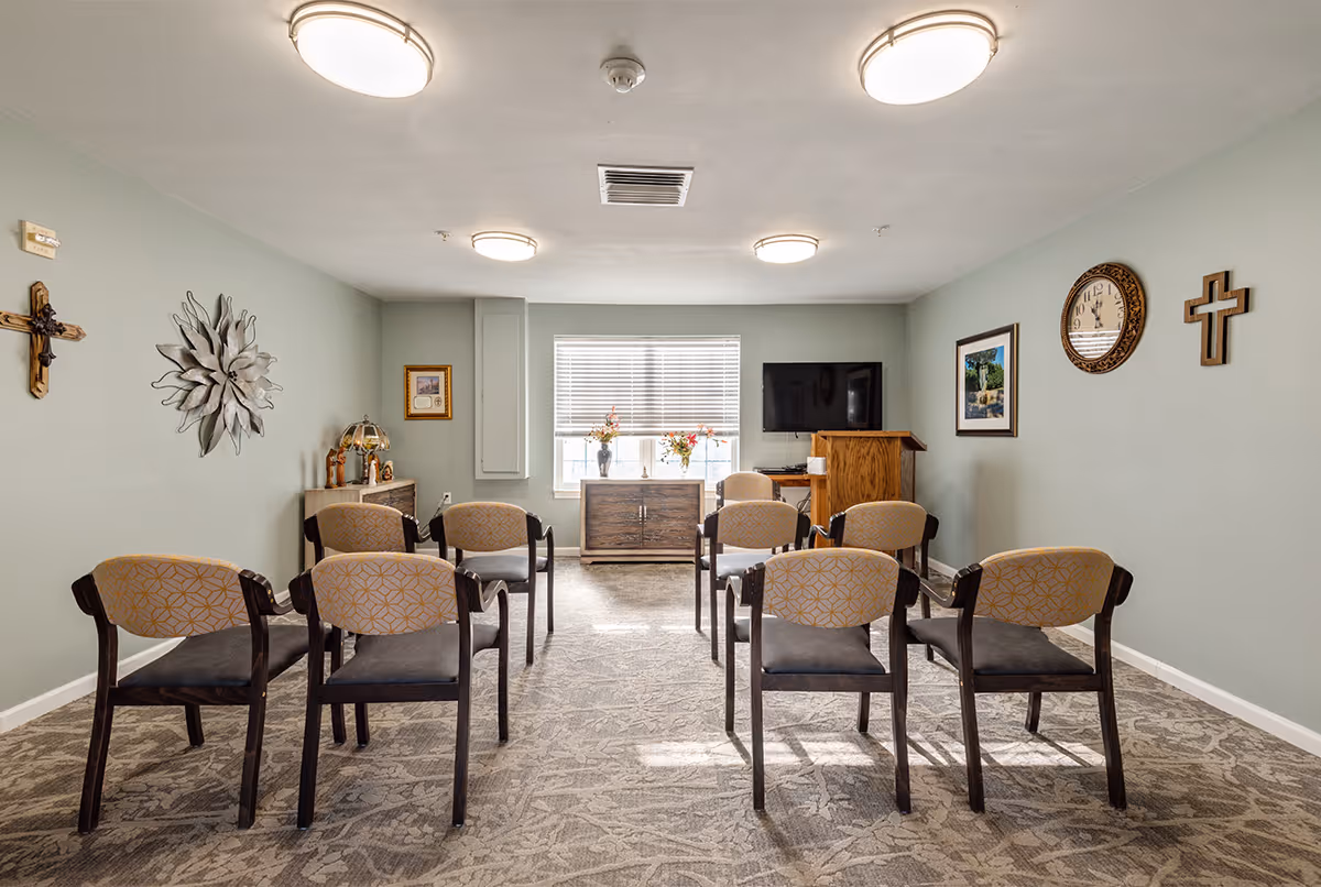 A small meeting or chapel room with eight chairs arranged in two rows facing a wooden podium and a wall-mounted TV. The room has light green walls decorated with two crosses, a metal flower wall art, a clock, and framed pictures. There is a window with blinds letting in natural light, and a cabinet with flower vases beneath it.