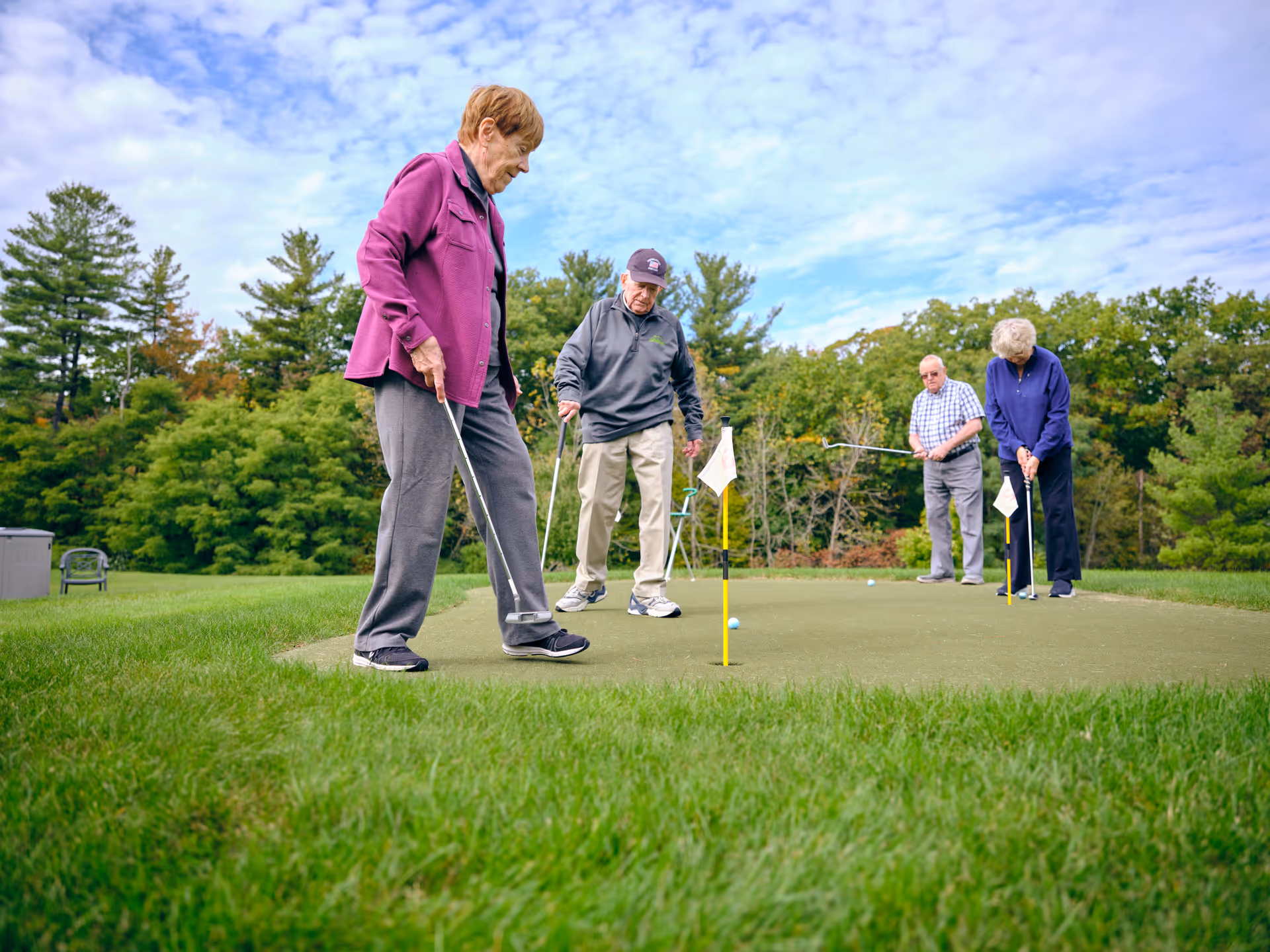 Four elderly people playing golf on a putting green outdoors surrounded by trees under a partly cloudy sky.