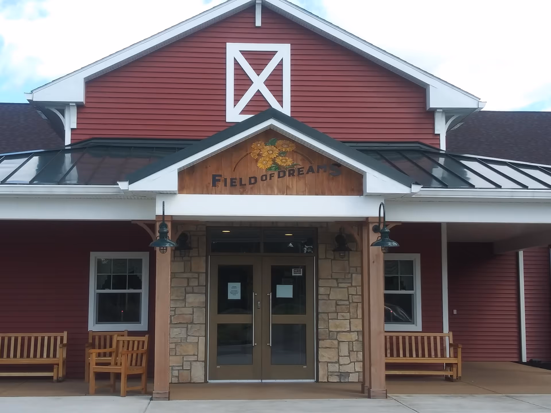 Front entrance of a building with red siding and white trim, featuring a sign above the door that reads 'Field of Dreams' with a sunflower design. There are two wooden benches on either side of the entrance and two green outdoor lamps hanging from the porch ceiling.