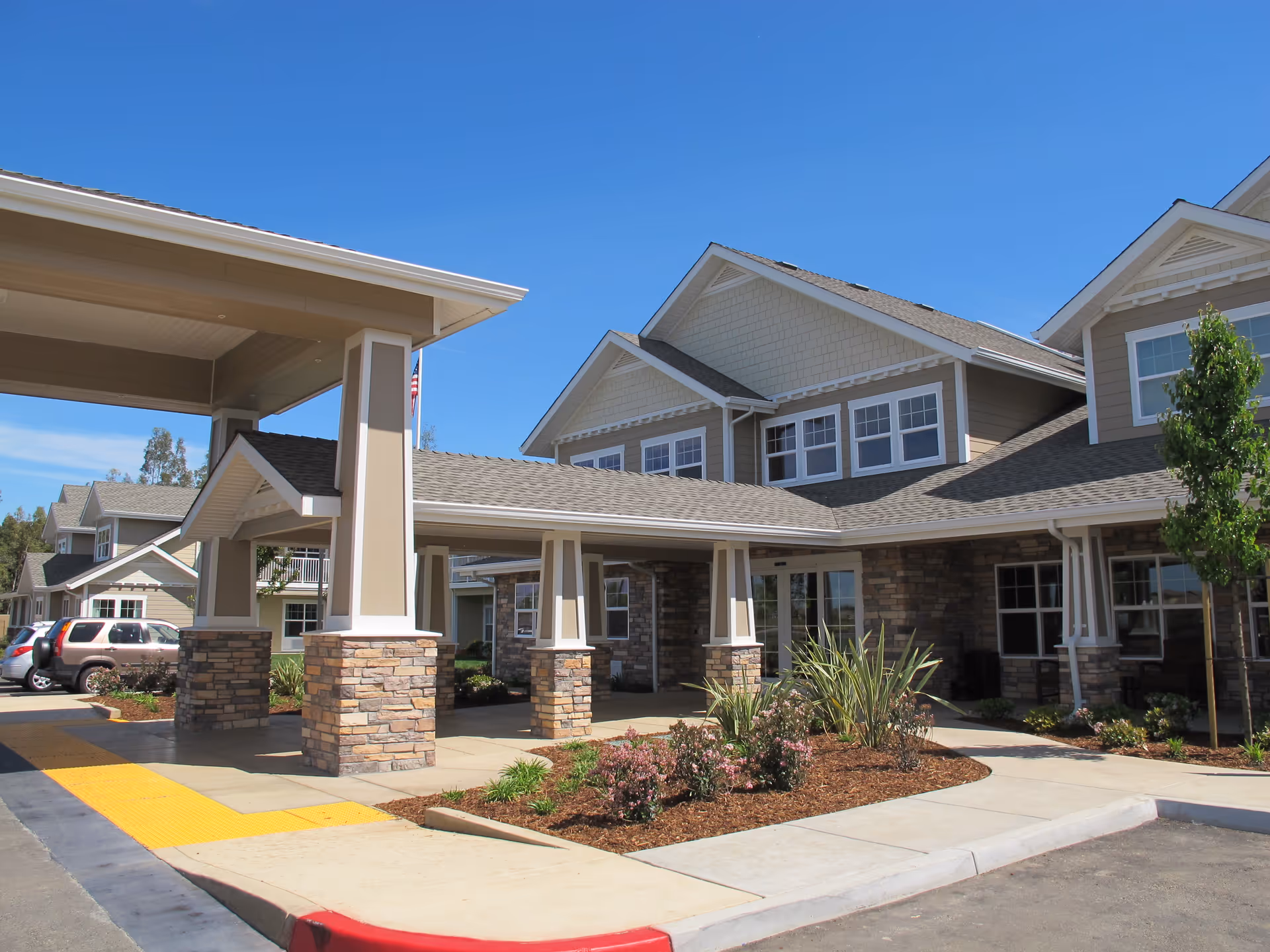 Exterior view of Meadowlark Estates Gracious Retirement Living showing a covered entrance with stone pillars, beige siding, multiple windows, landscaped plants, and a clear blue sky.