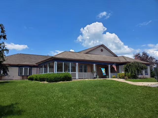 Exterior view of a single-story assisted living facility building with a covered porch, white railings, and an American flag near the entrance. The building is surrounded by a well-maintained green lawn and bushes under a blue sky with some clouds.
