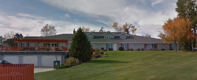 Low, single-story senior living building with a covered red porch, garages below, and a sloping green lawn under a partly cloudy sky.