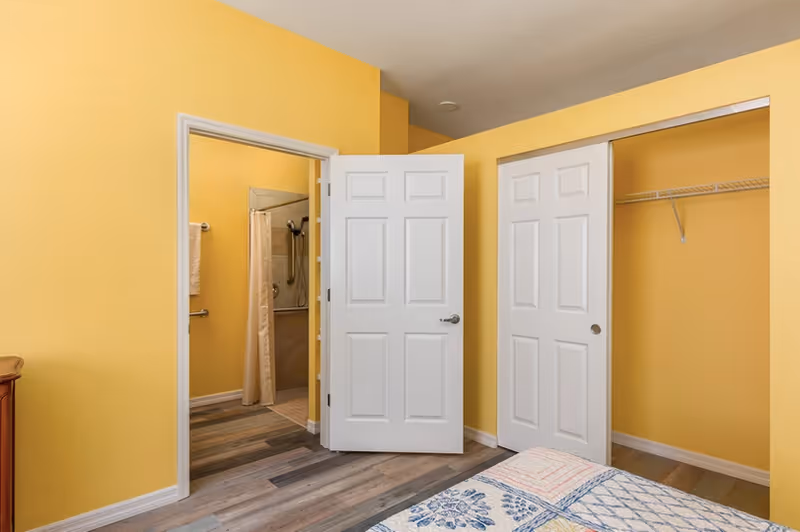 Interior view of a bedroom with yellow walls, a partially visible bed with a patterned quilt, an open closet with sliding doors, and an open door leading to a bathroom with a shower and grab bars.