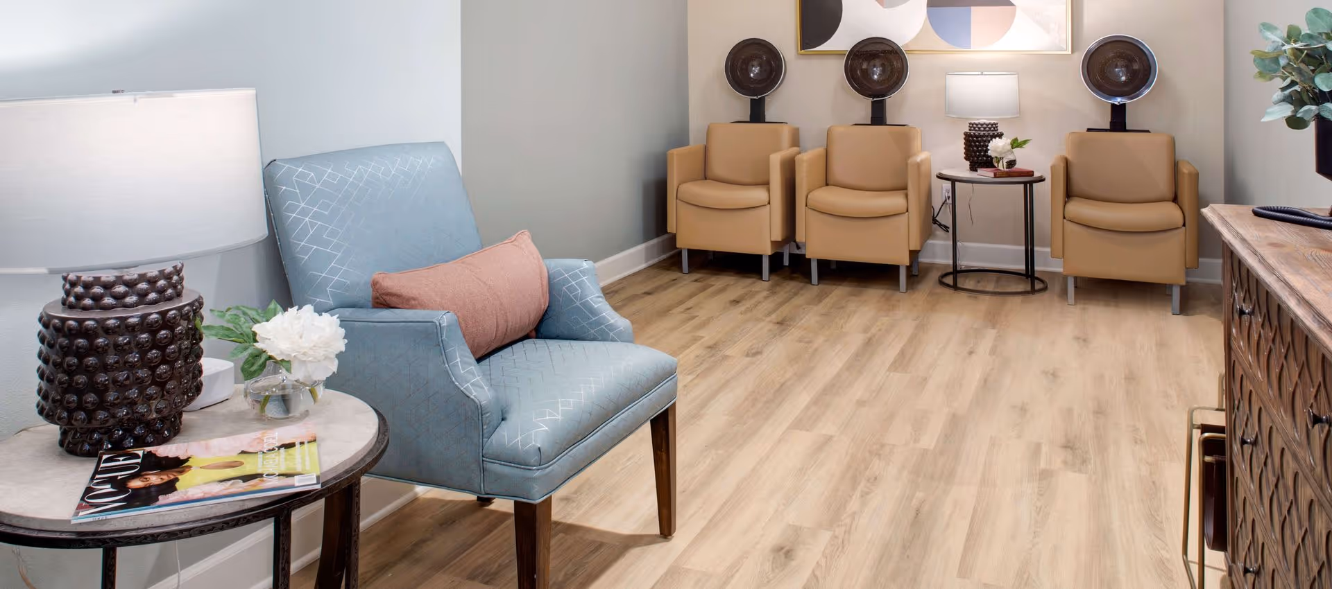 A bright seating area with a blue armchair and side table in the foreground and three tan chairs with hooded hairdryers along the back wall.