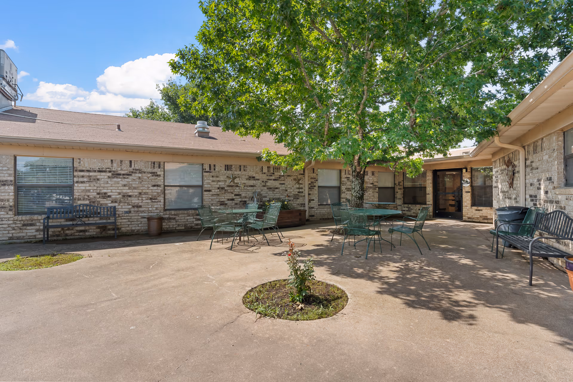 Outdoor courtyard area at Park Place Manor Rehabilitation and Healthcare featuring a large tree in the center, surrounded by metal tables and chairs, benches along the brick building walls, and a clear blue sky with some clouds.