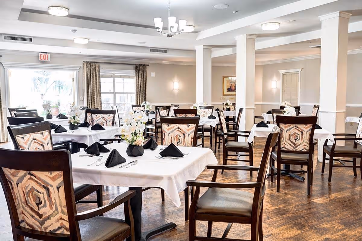 A bright and spacious dining room with multiple tables covered in white tablecloths, each set with black folded napkins, silverware, and floral centerpieces. The room features wooden chairs with patterned upholstery, hardwood floors, and soft lighting from ceiling fixtures and wall sconces. Large windows and glass doors allow natural light to fill the space.