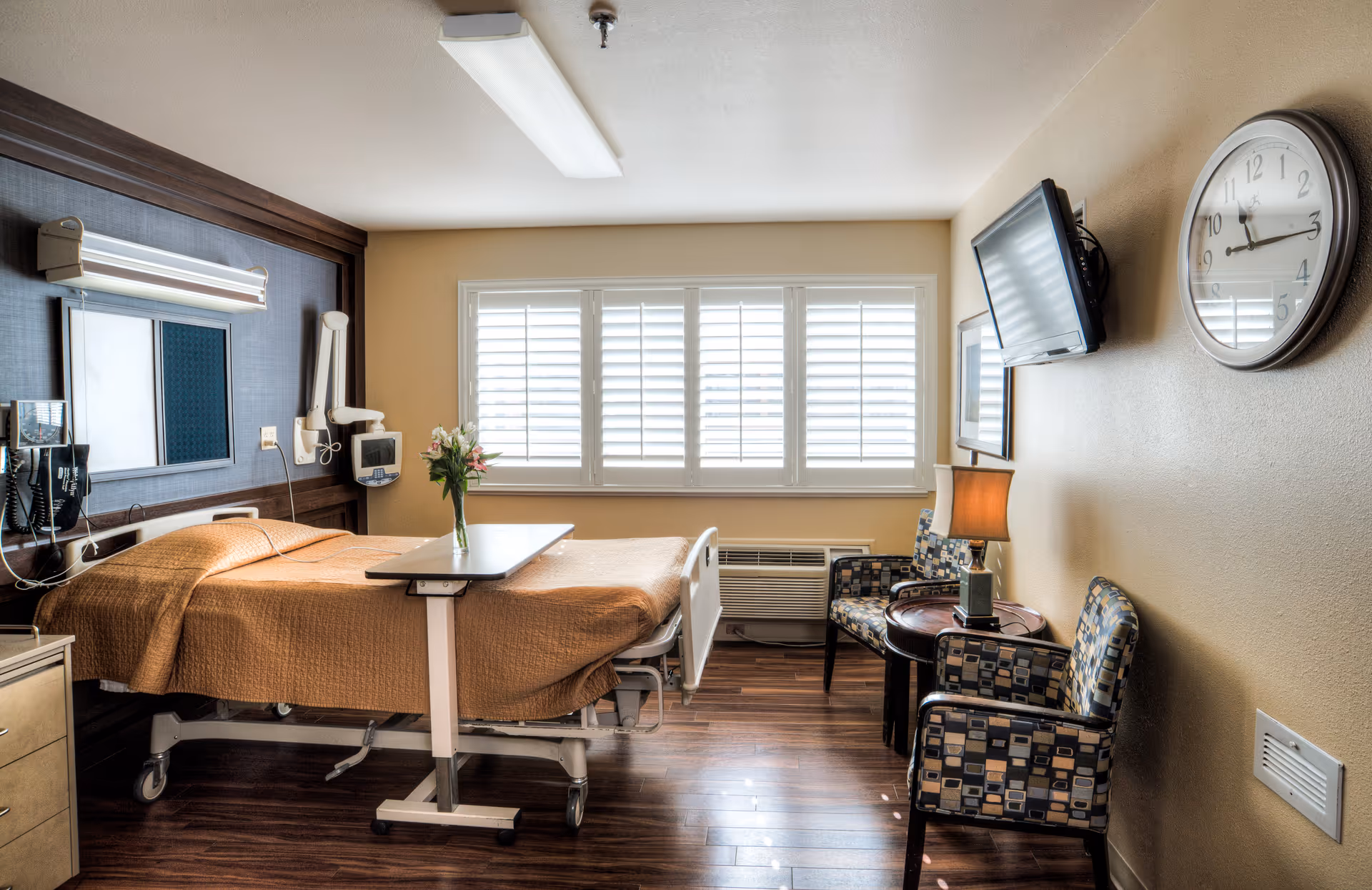 A well-lit healthcare facility room with a hospital bed covered in a brown blanket, a movable overbed table with a vase of flowers, two patterned armchairs with a small round table and lamp between them, a wall-mounted TV, a large clock, and a window with white shutters.