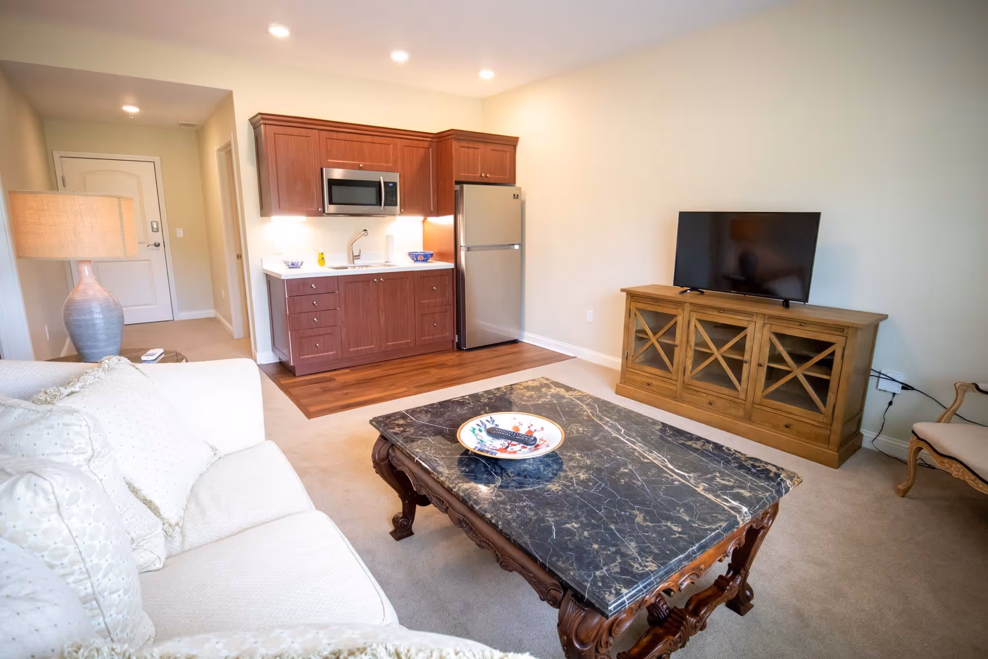 A cozy living area in a senior living facility featuring a white sofa with cushions, a marble-top coffee table with a decorative plate, a wooden TV stand with a flat-screen television, and a small kitchenette with wooden cabinets, a microwave, and a refrigerator. A lamp is placed on a side table next to the sofa, and there is a chair in the corner.