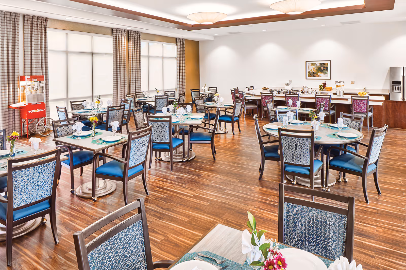A bright and spacious dining room with multiple tables set with plates, glasses, and napkins. The room features wooden floors, large windows with curtains, a popcorn machine in the corner, and a counter area with chairs and a refrigerator in the background.