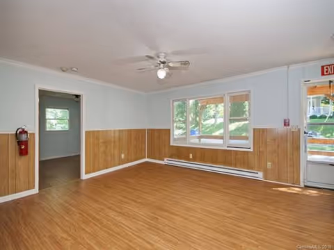 Empty senior living facility common area with wood paneling, large window, ceiling fan, exit door.