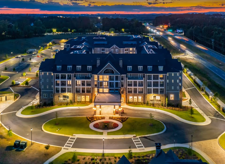Aerial view of a large, well-lit senior living facility named Holbrook Woodstock at dusk, with a circular driveway and landscaped grounds. The building has multiple floors with balconies and large windows, surrounded by parking lots and roads with light trails from passing vehicles.