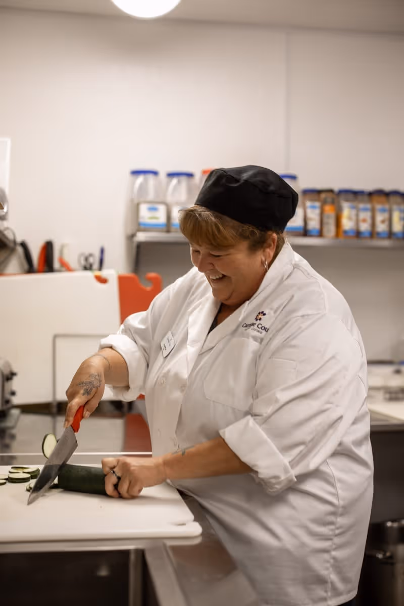 A smiling chef wearing a white coat and black hat slices a cucumber on a cutting board in a kitchen. Various containers of spices and ingredients are visible on a shelf in the background.
