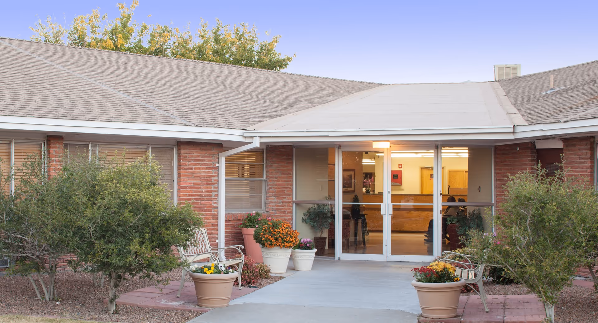 Entrance to Casa Del Sol Center, showing a single-story brick building with a covered walkway. There are glass double doors leading inside, flanked by potted plants and benches on either side. Small trees and shrubs are planted near the entrance.