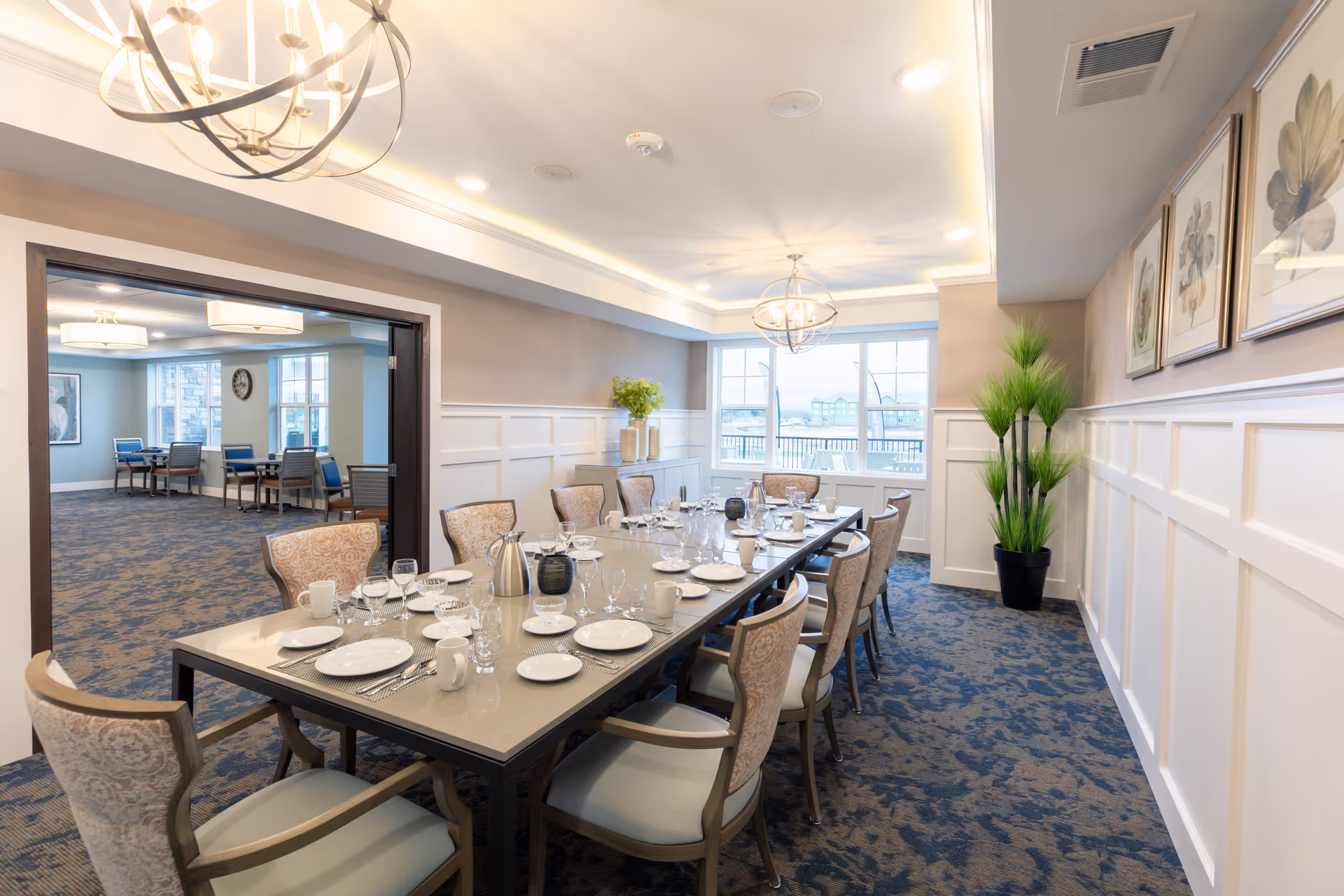 A well-lit dining room with a long rectangular table set for a meal, including plates, glasses, cups, and silverware. The room features patterned carpet, beige walls with white wainscoting, decorative framed artwork, and a large window providing natural light. There are upholstered chairs around the table and a potted plant in the corner. Adjacent room visible through an open doorway with additional tables and chairs.