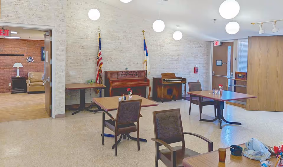 A senior living common/dining room with tables and chairs, two pianos against a white brick wall, flags, and round hanging lights.