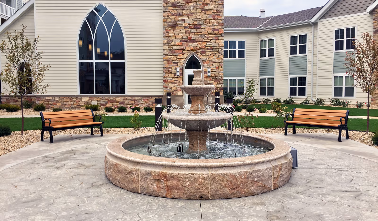 Outdoor courtyard area with a large stone water fountain in the center, flanked by two wooden benches with black metal frames. The background shows a building with beige siding, stone accents, and large arched windows, surrounded by small trees and landscaping.