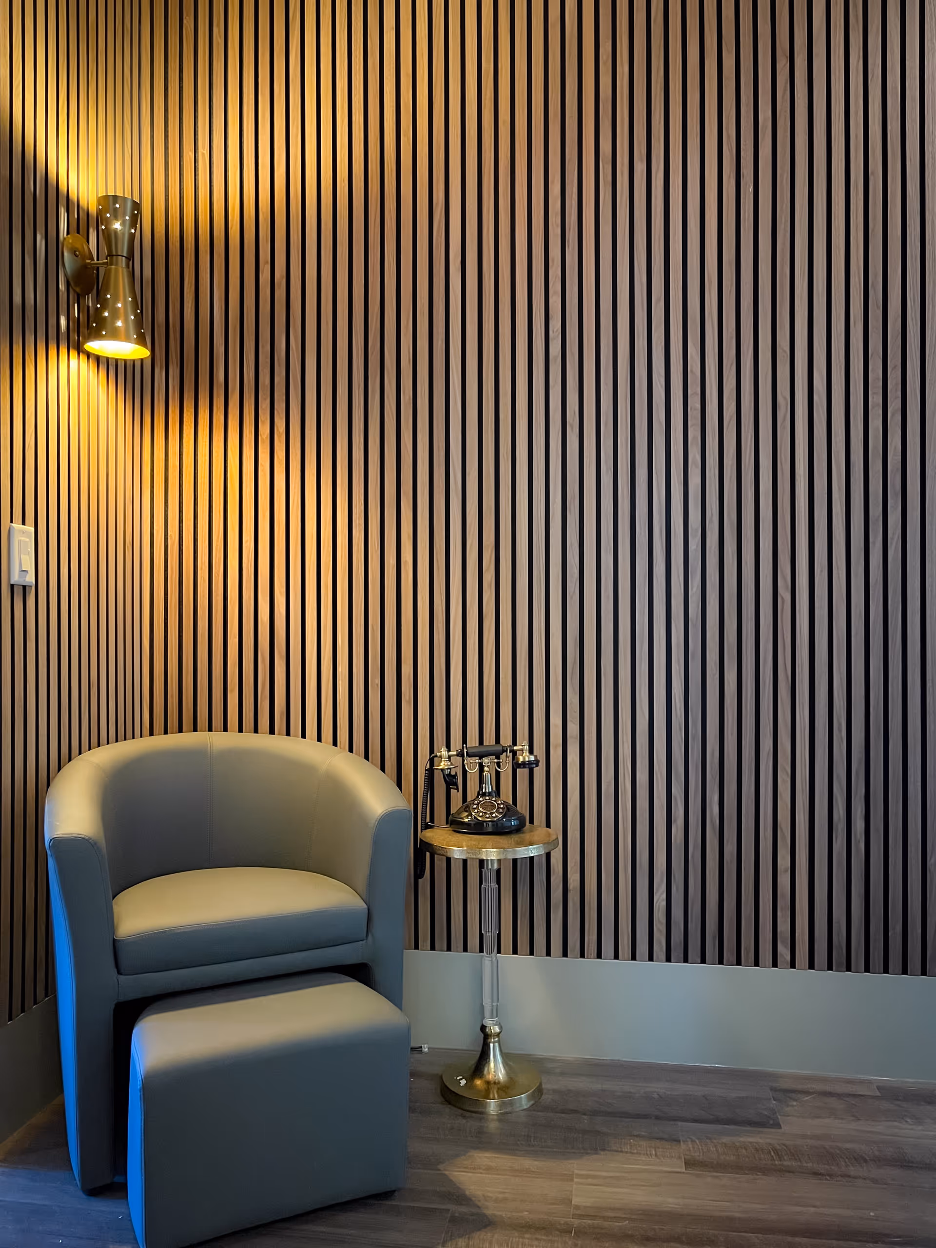 A cozy corner with a gray upholstered armchair and matching ottoman on a wooden floor. Next to the chair is a small round side table with a vintage black rotary telephone. The background features a modern wooden slat wall with a brass wall sconce light fixture casting a warm glow.
