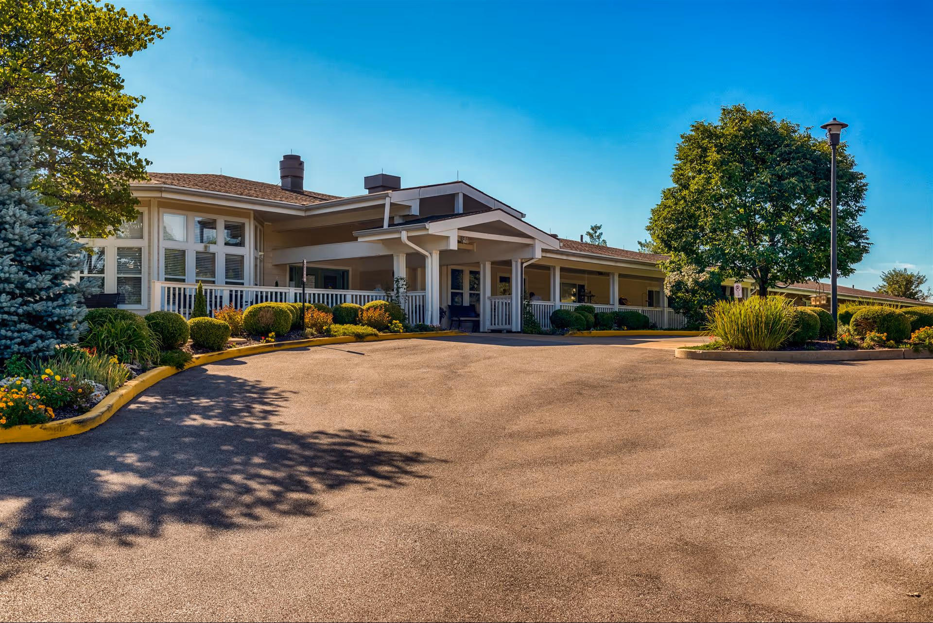 Front entrance of a single-story assisted living facility with a covered porch, landscaped beds, and a circular driveway under a clear blue sky.