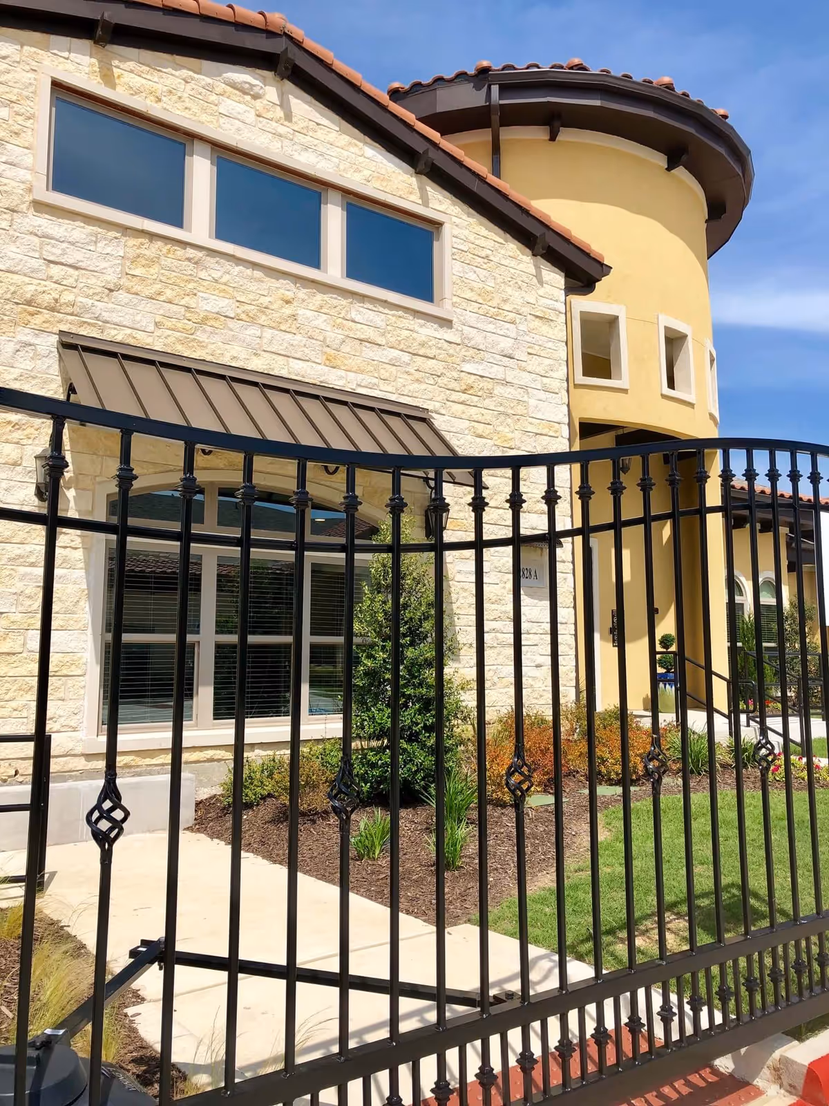 Exterior view of a senior living facility with a stone and stucco facade, featuring large windows and a decorative black metal fence in the foreground. The building has a tiled roof and landscaped greenery around the entrance.
