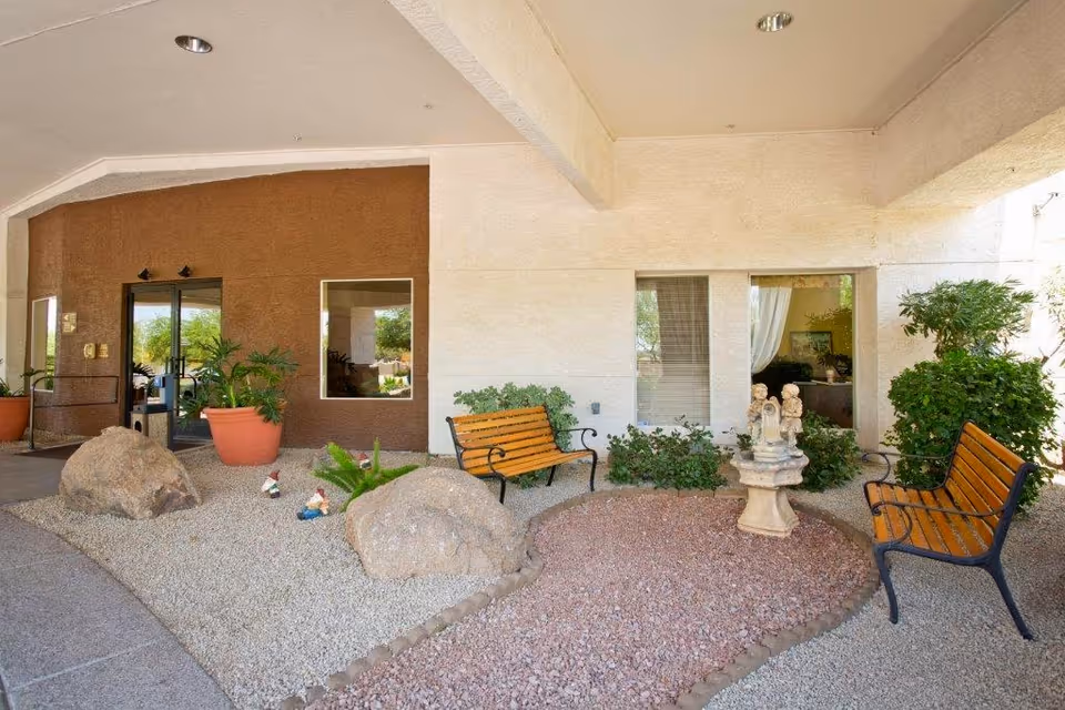 Covered outdoor seating area at Discovery Point Retirement Community featuring two wooden benches with black metal frames, a small decorative stone fountain with cherub statues, large rocks, potted plants, and gravel landscaping under a beige ceiling.