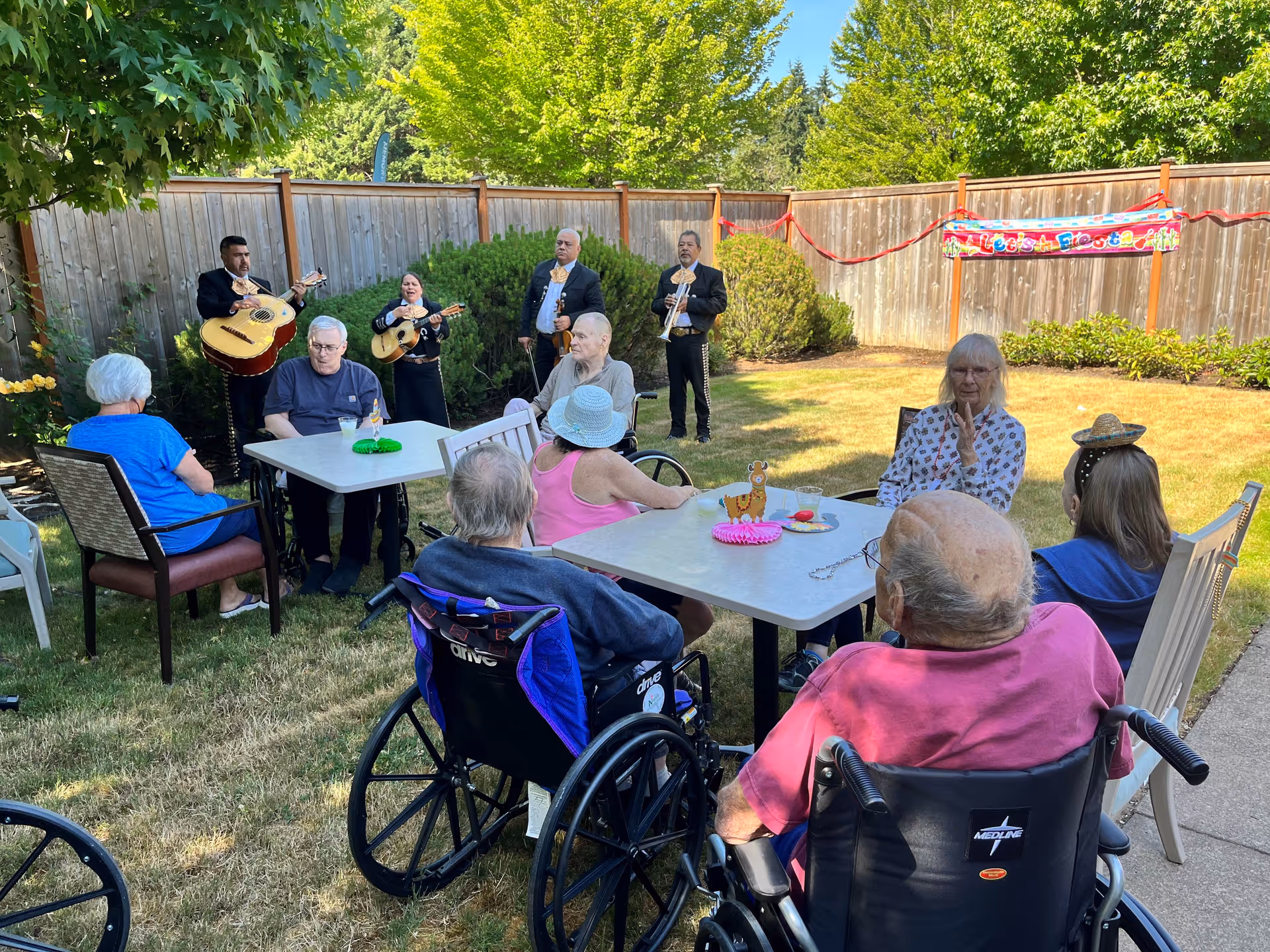 A group of elderly people sitting outdoors in a fenced yard, some in wheelchairs and others in chairs, gathered around two tables with drinks and decorations. In the background, a mariachi band with three musicians is playing instruments. There is a colorful banner on the wooden fence that reads 'Let's Fiesta'.