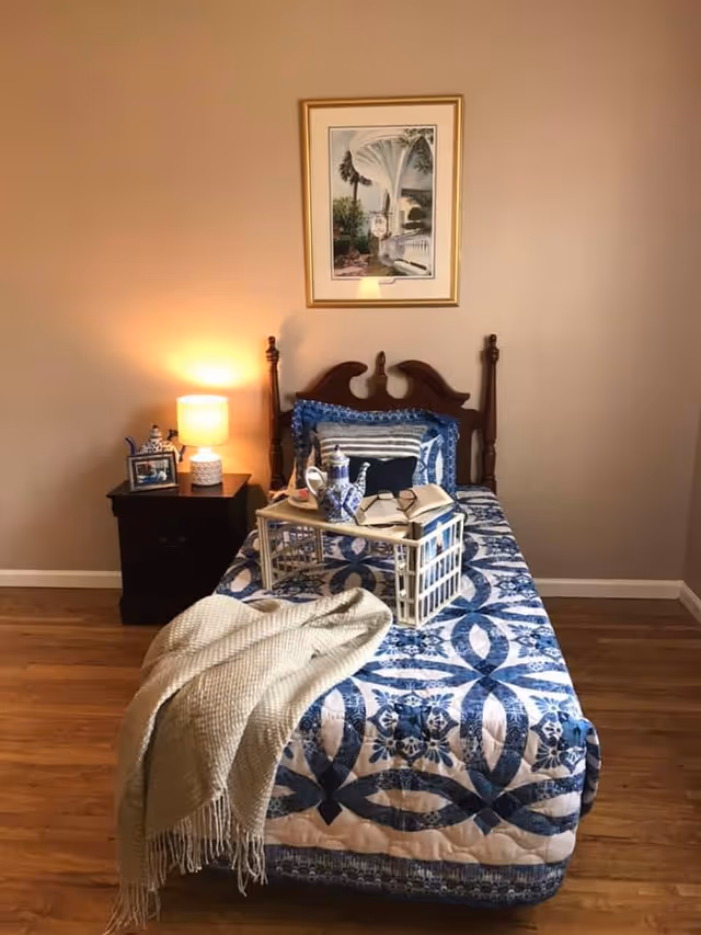 A single bed with a blue and white patterned quilt and multiple pillows is positioned against a beige wall. On the bed is a wooden tray holding a teapot, cup, and an open book. A beige throw blanket is draped over the foot of the bed. To the left of the bed is a dark wooden nightstand with a lit lamp, a small framed photo, and a decorative item. Above the bed hangs a framed picture of an outdoor scene with columns and greenery. The floor is wooden.