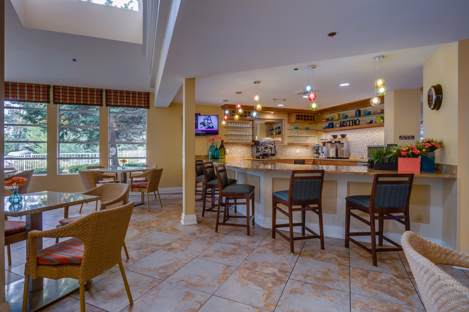 A bright and cozy bistro area with a counter and several high chairs. The counter has coffee machines and shelves with cups and decorative items. There are multiple small tables with wicker chairs featuring red and green striped cushions. Large windows with plaid valances let in natural light and offer a view of greenery outside. Colorful pendant lights hang from the ceiling, adding a warm ambiance.