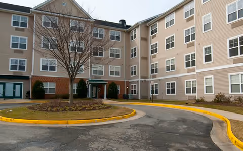 Exterior view of a multi-story senior living facility building with beige siding and brick accents. There is a circular driveway with a landscaped island featuring a leafless tree and shrubs in front of the entrance.