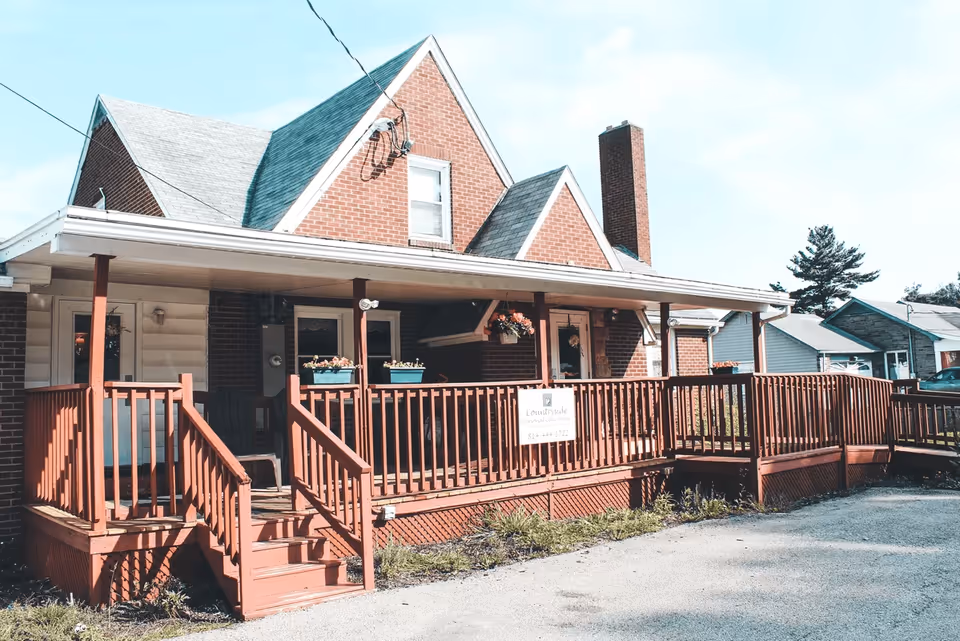 Exterior view of a brick and siding building with a covered wooden porch and ramp. The porch has flower pots and a sign that reads 'Countryside Personal Care Home'. The building has a steep roof with multiple gables and a chimney. There are neighboring houses and a clear sky in the background.