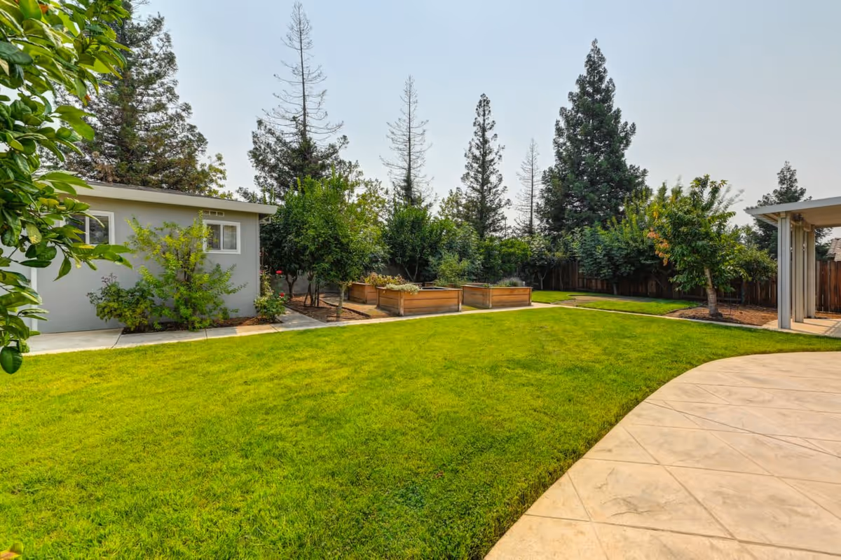 Well-maintained grassy backyard with raised garden beds, trees, a small outbuilding, and a curved concrete patio.