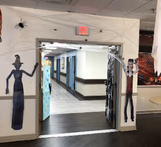 Interior view of a hallway in a senior living facility decorated with Halloween-themed cutouts including a witch, a vampire, and large spiders on the walls around a doorway. The hallway has light-colored walls with dark trim and a light wood floor. An exit sign is visible above the doorway.