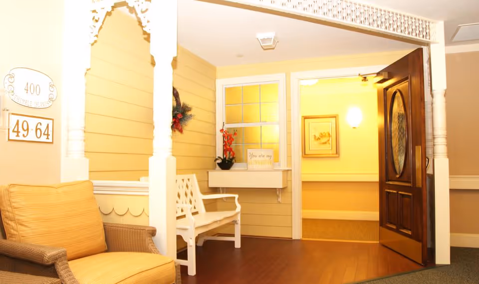 A cozy indoor seating area with a cushioned wicker chair and a white wooden bench. The walls are painted yellow with white trim and decorative molding. There is a dark wooden door with an oval glass panel open to a warmly lit hallway. A small window with a flower pot and a sign that reads 'You are my SUNSHINE' is visible. Room number signs 400 and 49-64 are mounted on the wall.