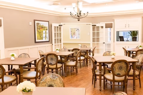 A bright dining room with several wooden tables and upholstered chairs arranged neatly. The room features hardwood floors, cream-colored walls with wainscoting, a chandelier hanging from the ceiling, and framed artwork on the wall. Small flower arrangements are placed on each table. French doors and a window with a view into another room are visible in the background.