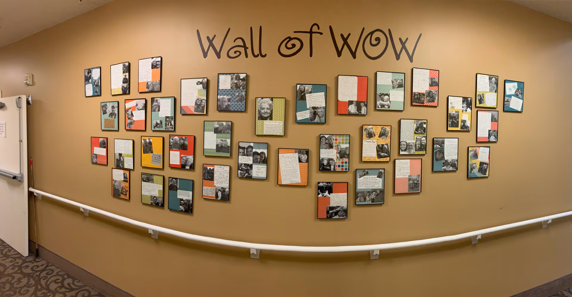 A hallway wall display titled 'Wall of WOW' featuring multiple framed notes and photographs arranged in a grid pattern. The wall is painted beige and has a white handrail running along its length. The display appears to showcase positive messages or testimonials with pictures of people.