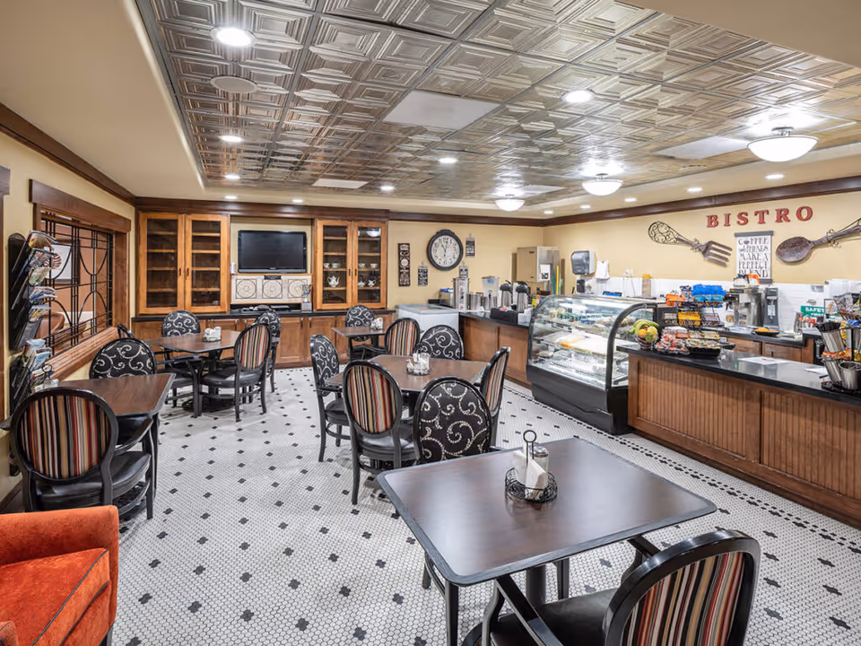 Interior view of a bistro area in a senior living facility with multiple tables and chairs arranged on a tiled floor. The ceiling has a decorative pattern with recessed lighting. On the right side, there is a counter with a glass display case containing food items, coffee machines, and snacks. The back wall has a clock, cabinets, and a sign that reads 'BISTRO' with decorative utensils.