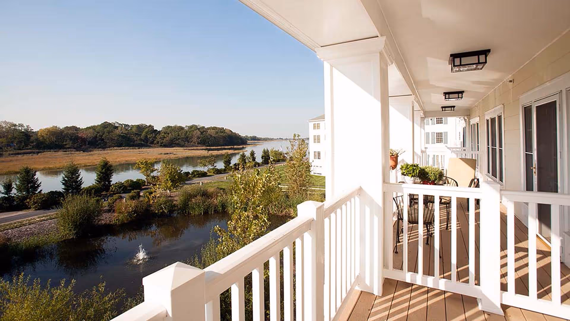 View from a covered balcony with white railings overlooking a small pond with a fountain, landscaped greenery, and a river with trees in the background under a clear sky.