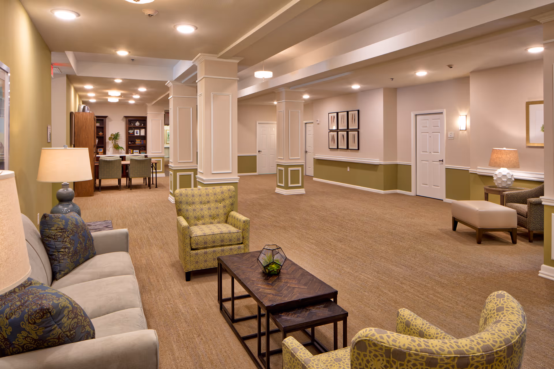 A spacious and well-lit common area in an assisted living facility featuring comfortable seating including a sofa and armchairs arranged around a wooden coffee table. The room has beige walls with green accents, decorative columns, framed artwork, and a dining area with a table and chairs in the background.