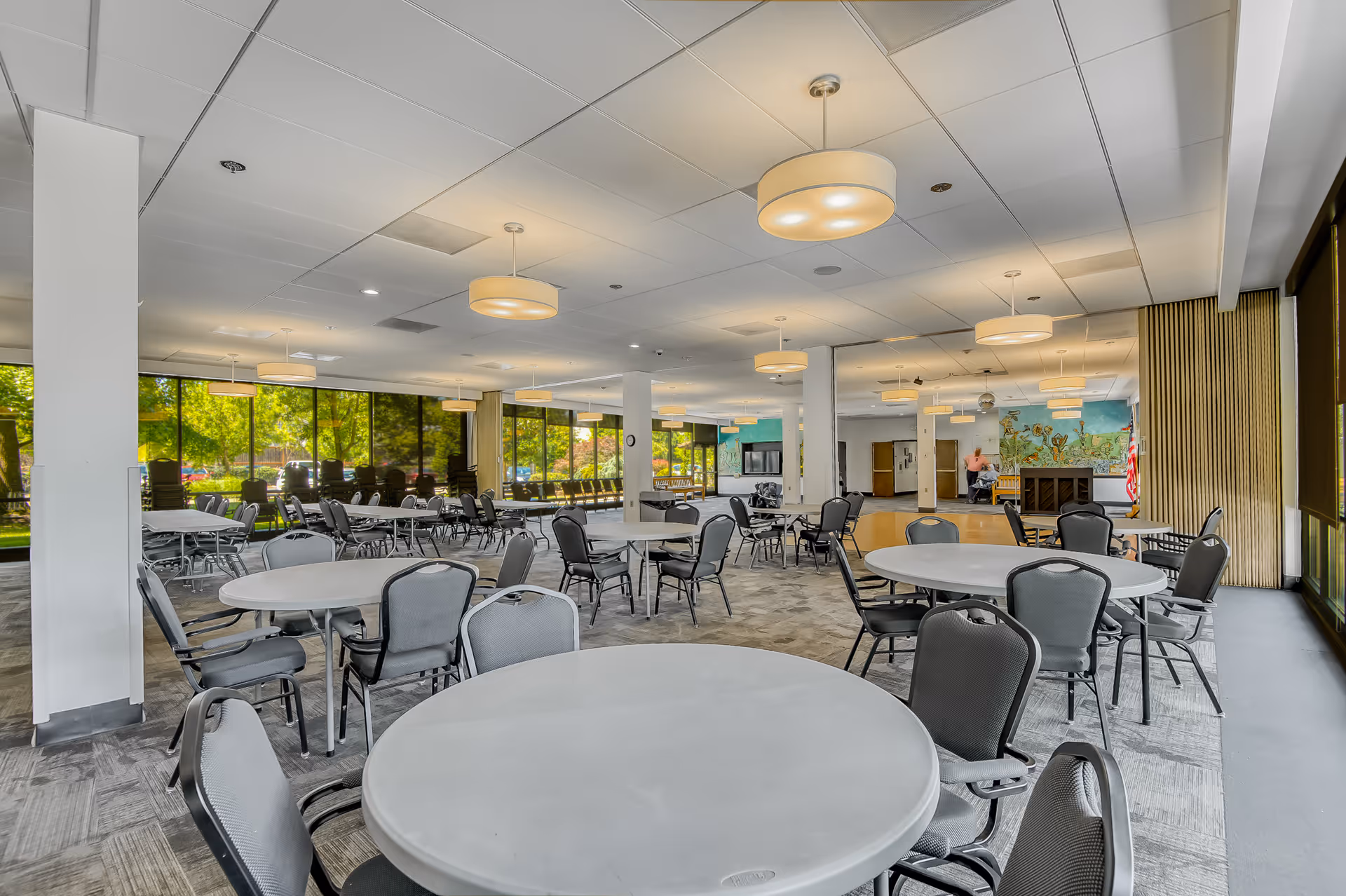A spacious dining room with multiple round tables and gray chairs arranged neatly. Large windows on one side let in natural light and offer a view of green trees outside. The ceiling has several round pendant lights, and the floor is carpeted. In the background, there is a mural on the wall and a person standing near a piano.